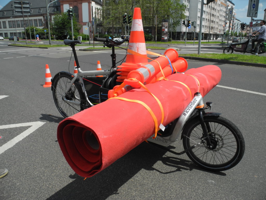 Ein Lastenrad, voll beladen mit Verkehrspylonen und einem breiten roten Teppich.